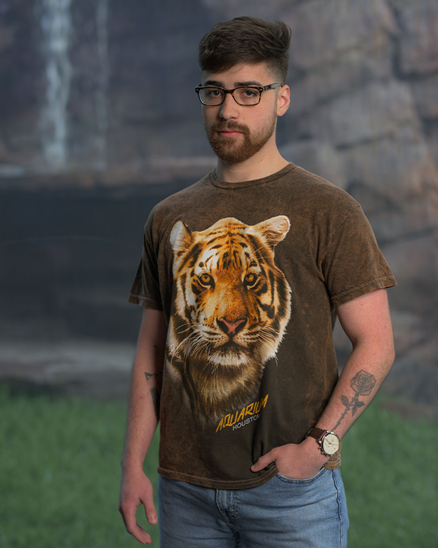 A person is seen wearing a brown t-shirt with a strikingly realistic print of a tiger’s face, below which the text ‘AQUARIUM HOUSTON’ is printed. They are standing in front of a scenic backdrop featuring rocks and a waterfall, enhancing the naturalistic theme. They are also wearing blue jeans, a watch, and have tattoos on their left arm.