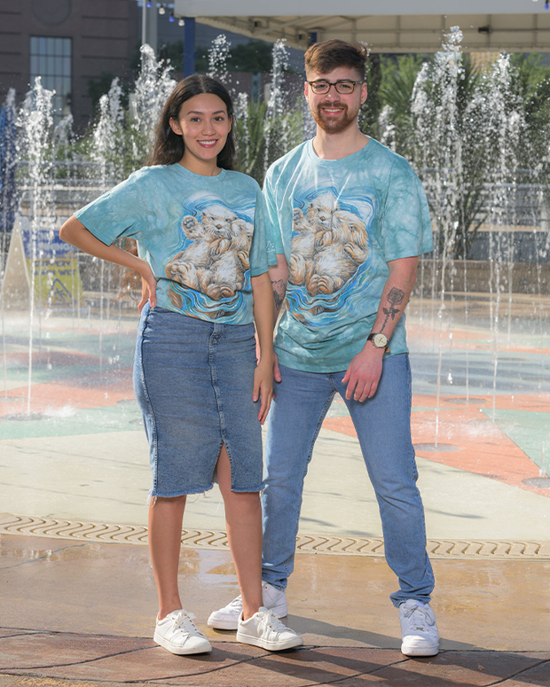 Two individuals are standing side by side in front of a water fountain, wearing matching blue tie-dye t-shirts featuring a two otters design. One person is dressed in a denim skirt and the other in light blue jeans, both wearing white sneakers. The fountain in the background adds a dynamic element to the scene, with water jets reaching upwards, and a building structure is faintly visible in the distance.