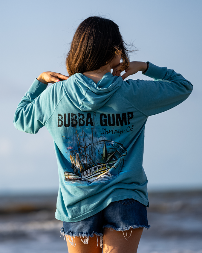 A knee-up view of a brunette model standing in front of the sea and the sky, with her hands touching the top of her blue hoodie featuring a graphic of a boat at sea with compass design behind it, reading "Bubba Gump Shrimp Co." above it. She is also wearing blue jean shorts.