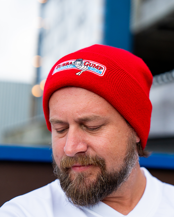 A headshot of a model in a white V-neck shirt wearing a red beanie with an embroidered patch on the front featuring a cartoon shrimp character in a top hat and text that reads "BUBBA GUMP SHRIMP CO." The background includes an out-of-focus building and lights.
