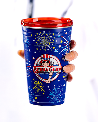 The image you uploaded depicts a hand holding a vibrant blue cup. The cup has a bright red rim at the top and is adorned with white stars and fireworks designs, giving it a festive appearance. In the center of the cup, there’s a logo that reads “BUBBA GUMP SHRIMP CO.” Above the logo, there’s an image of a shrimp wearing sunglasses and an American flag-themed hat. The background is plain white, making the colorful cup stand out prominently.