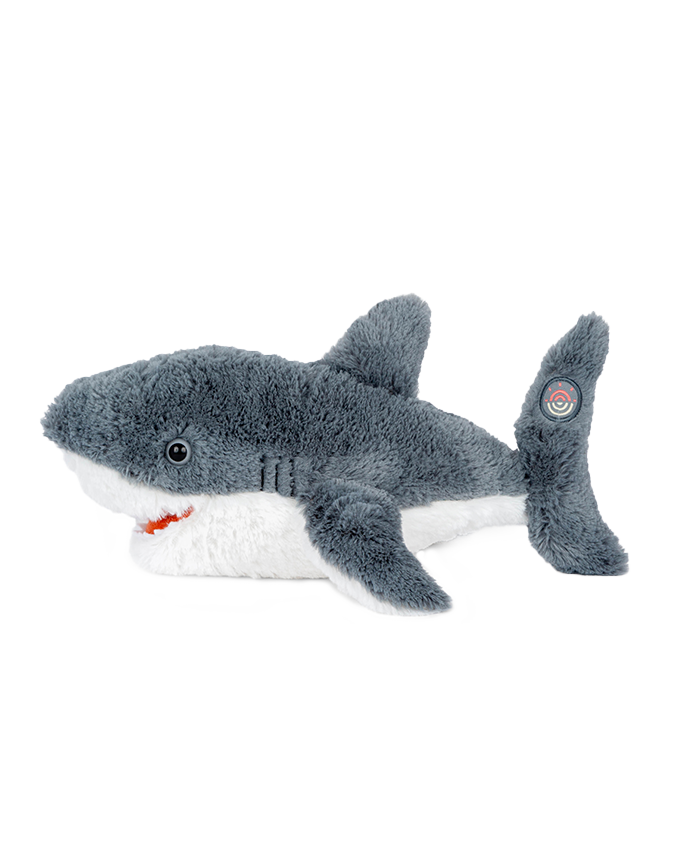 A side view of a plush grey shark toy with a white underbelly and an open mouth, featuring a circular target label on the tip of the tail. The background is plain white.