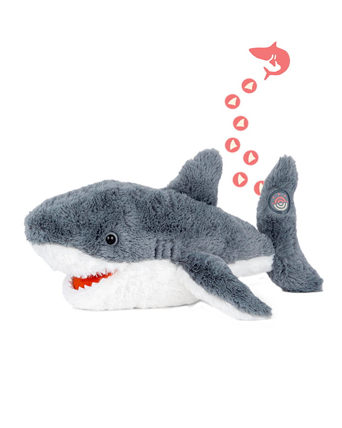 A grey shark plush toy with a white underbelly and an open mouth. There is an illustration above the shark depicting a smaller shark icon with several small red hearts trailing behind it, leading to a target symbol on the side fin of the plush toy. The background is plain white.
