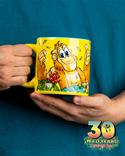 A commemorative yellow mug with a colorful illustration of a cheerful orangutang surrounded by foliageand is waving, set against a teal shirt. The mug celebrates ‘30 WILD YEARS!’ at the Rainforest Cafe.