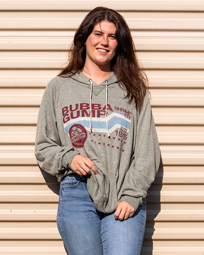 Brunette model leaning against wall wearing army green bubba gump hoddie and blue jeans. Green hoodie has drawstrings, in burgundy letters "bubba gump shrimp co.", on top. Below is a rectangular design with white lines and a thick light blue line in the center. On bottom right, a burgundy bubba gump logo, next to it, two lines of burgundy stars.
