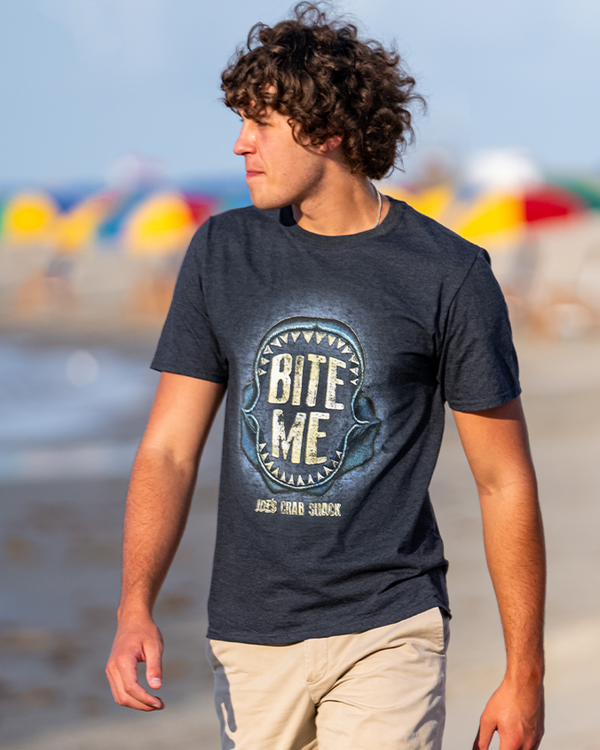 A person standing on a sunny beach full of umbrellas, wearing a dark crewneck t-shirt with an open shark jaw skull with text reading "BITE ME" inside of it, and "JOE'S CRAB SHACK" under it, paired with khaki shorts.
