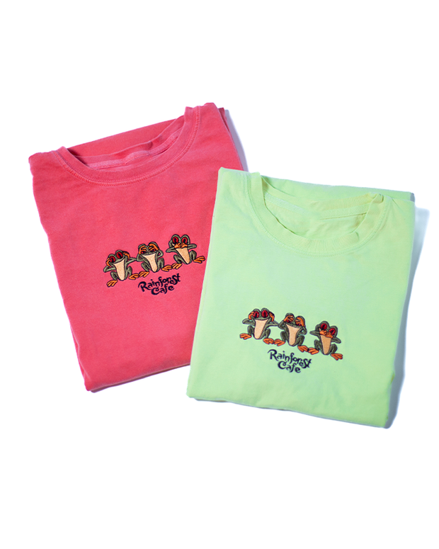 Two neatly folded t-shirts, one pink and one light green, each featuring an embroidered design of a group of colorful frogs with ‘Rainforest Cafe’ written underneath. The frogs are depicted in various poses, adding a playful touch to the shirts. They are placed side by side against a white background, with shadows indicating a light source from above.