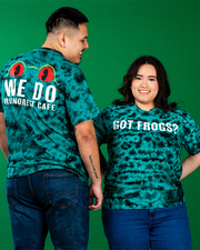 Two people stand side by side against a green background, both wearing tie-dye t-shirts. The person on the left has a shirt that reads ‘WE DO RAINFOREST CAFE’ with two red eyes above the text, and the person on the right has a shirt with the phrase ‘GOT FROGS?’ Both are wearing denim bottoms.