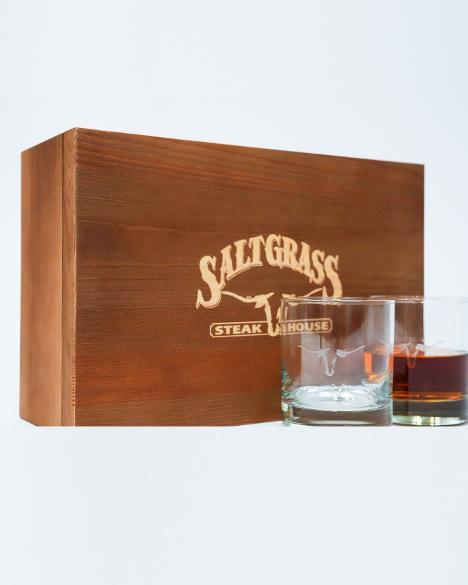 A wooden box with "Saltgrass Steak House" printed on it in front of two whiskey glasses with the Saltgrass Longhorn Head in the center, one filled with liquid. The background is plain white.