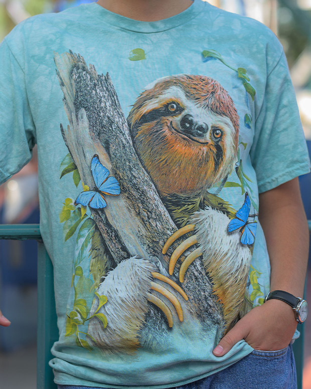 A person is seen wearing a light blue t-shirt that showcases a detailed and colorful print of a sloth clinging to a tree branch. The sloth, depicted in shades of brown and white, appears relaxed and content. The design is accentuated with vibrant blue butterflies and green leaves, creating a lively and naturalistic theme.