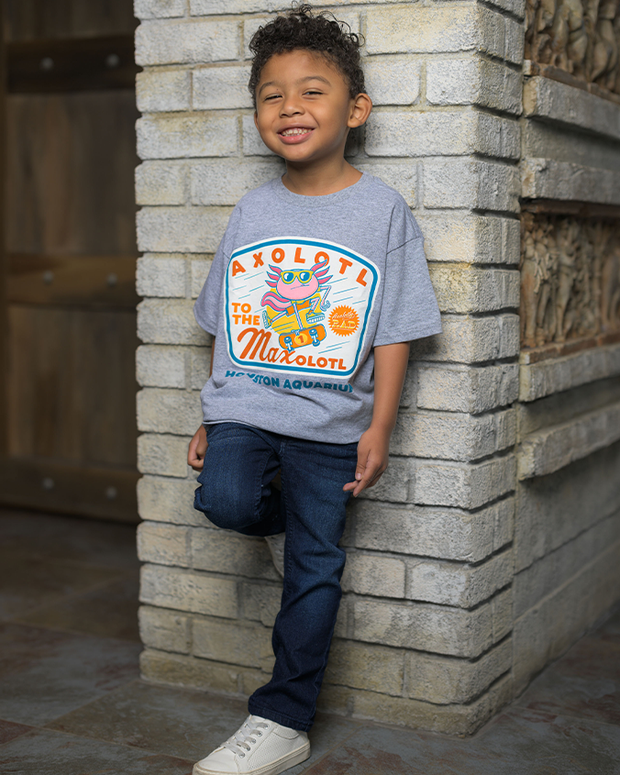 A kid is seen leaning against a textured stone wall, wearing a grey t-shirt with a vibrant axolotl design and the playful words ‘AXOLOTL TO THE MAXOLOTL HOUSTON AQUARIUM’. The casual outfit is completed with dark jeans and white sneakers. To the side, a wooden door adds a rustic touch to the setting.