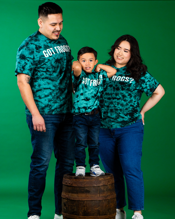 Two parents and a little boy standing against a green background, the green boy on a barrel. The group is wearing matching outfits including blue jeans, white sneakers, and green and black tie-die T-shirts that say, "GOT FROGS?" in the center in bold text.