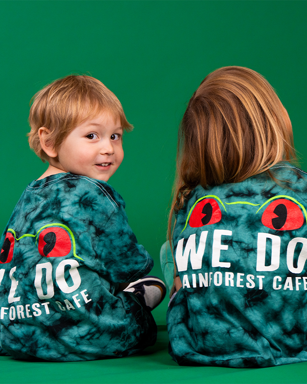 A backside view of little kids sitting against a green backdrop, wearing a green and black tie-dye shirt with a graphic of red frog eyes, and white bold text reading "WE DO RAINFROEST CAFE".