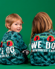 Two individuals are seen from the back, wearing black and green tie-dye shirts with a circular pattern. The shirts features the same frog encircled by the text ‘WE DO IT IN THE RAINFOREST CAFÉ.’ Both are set against a solid green background