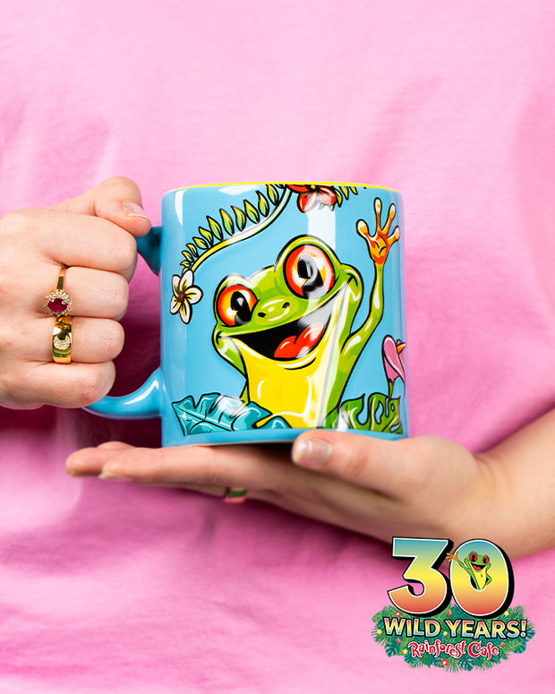 A person holding a vibrant blue mug with a colorful illustration of Cha Cha the frog from Rainforest Cafe. The mug is adorned with tropical leaves and flowers, and the text ‘30 WILD YEARS! Rainforest Cafe’ is visible. The background is pink, complementing the playful and whimsical design of the mug.