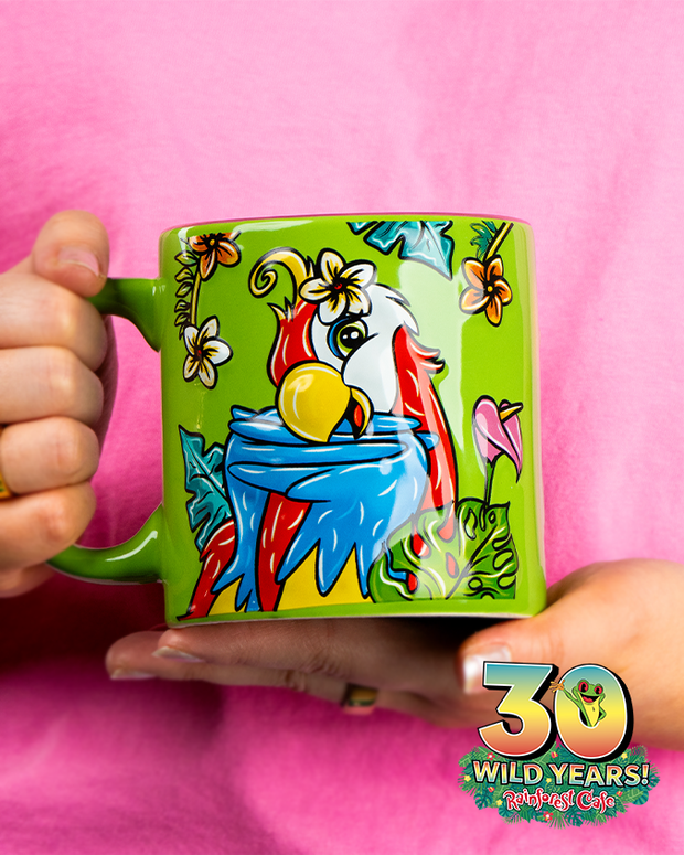 A vibrant green mug adorned with a colorful illustration of a parrot surrounded by tropical flowers, held against a pink shirt. The mug features an emblem celebrating ‘30 WILD YEARS!’ of Rainforest Cafe with Cha Cha Sticking out the 0.