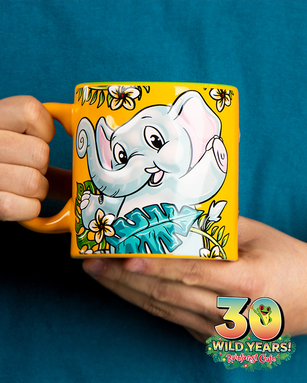 A colorful mug with a vibrant illustration of a smiling elephant surrounded by tropical foliage, held in someone’s hands against a teal shirt. The mug features a bright yellow body with an orange handle and an emblem celebrating ‘30 WILD YEARS!’ of Rainforest Cafe.