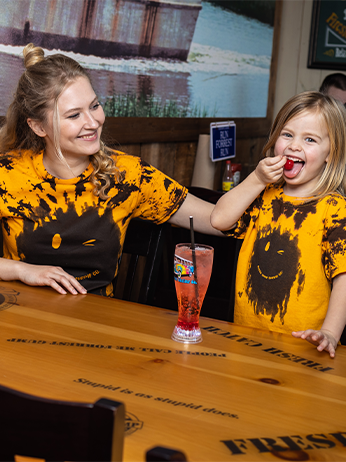 The image depicts two individuals seated at a wooden table. They are wearing matching yellow t-shirts with black ink splatter patterns and smiley faces. In front of one individual is a tall glass of pink beverage with a straw. The background features a rustic wall with framed artwork, and there’s a small blue sign visible.