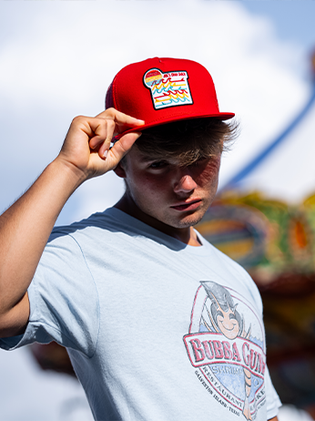 An individual wearing a red cap with a logo and a gray t-shirt with an illustration and text, standing against a blurred outdoor background. The cap’s logo appears colorful and cartoonish, while the t-shirt graphic includes a character and text that are not fully legible due to the angle and folds of the fabric.