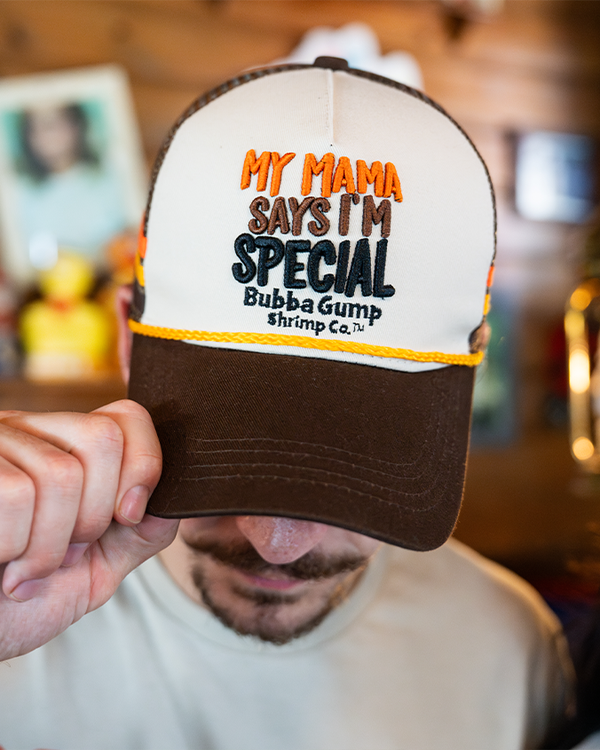 A close-up view of a model in a white crewneck T-shirts holding onto a brown and white baseball cap he's wearing, which says "MY MAMA SAYS I'M SPECIAL BUBBA GUMP SHRIMP CO." in the center in multi-color. The background is a blurry log cabin setting.