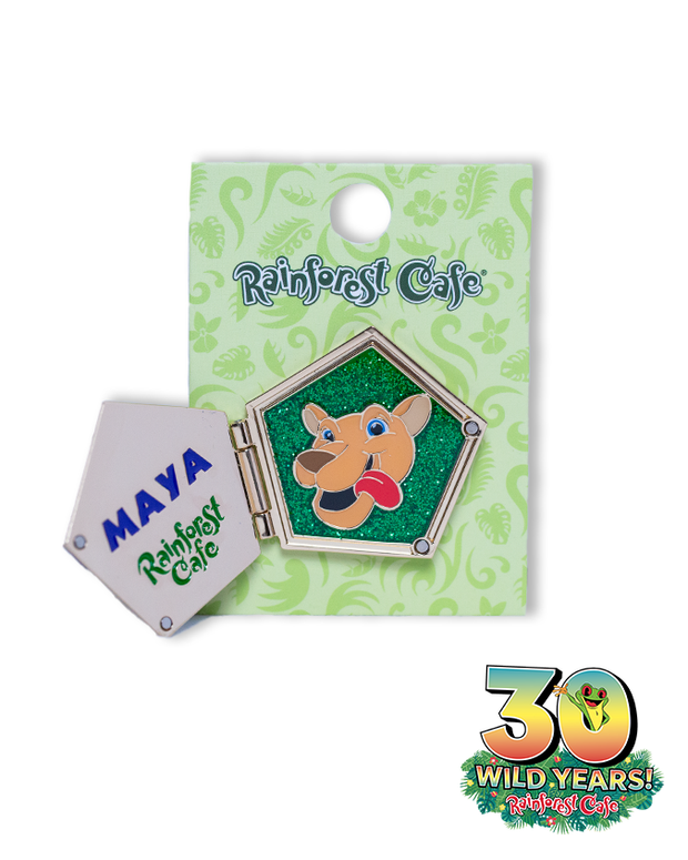 A collectible pin from Rainforest Cafe. Left side feauture the name ‘MAYA’ in blue letters, under it in green 'RAINFOREST CAFE'. Right side has animated character against a sparkly green background encased in a hexagonal frame, presented on a decorative card. Bottom right corner has rainforest cafe’s 30th-anniversary logo with a green frog coming out the 0.