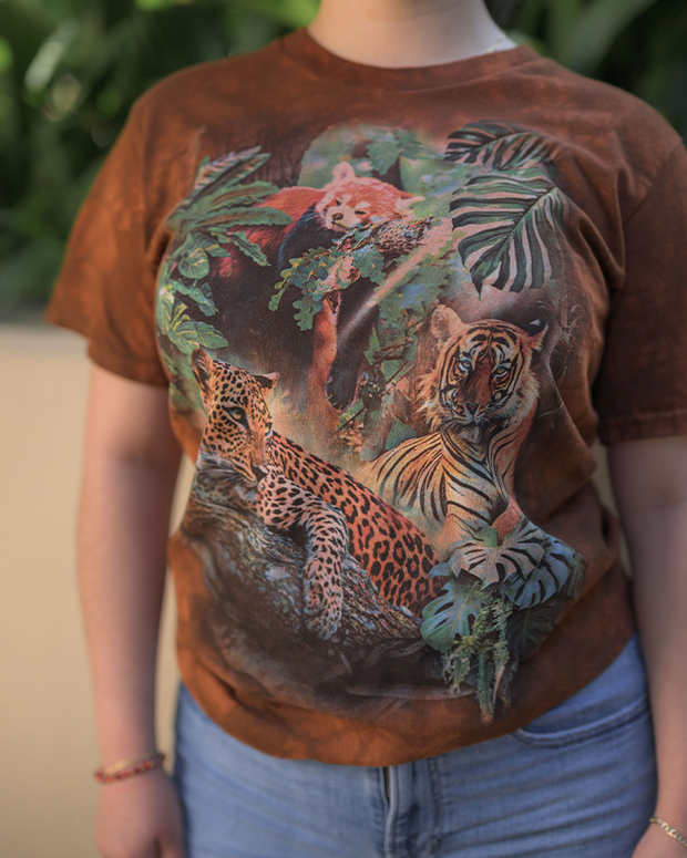 A person is seen wearing a brown t-shirt with an intricate and colorful design. The design on the t-shirt depicts a vibrant jungle scene featuring a tiger and leopard amidst lush green foliage. The tiger is positioned towards the bottom right, while the leopard is towards the bottom left of the design. Above them, there's a red panda amidst the leaves. The person is wearing blue jeans, which are partially visible in the image. They are also wearing bracelets on their left wrist.