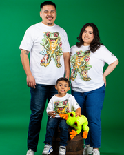 Three individuals stand against a green backdrop, each wearing white t-shirts with colorful frog designs. The tallest person on the left sports a tattoo on their arm. In the center, a child sits on a wooden barrel, clutching an orange and green plush frog toy. The person on the right places a hand on their hip. Their casual jeans and sneakers complement the relaxed and cheerful scene.