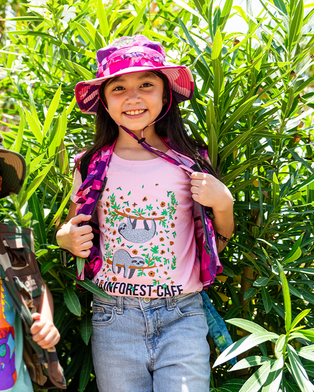 little girl in front of plant bush, wearing Pink tee with two cartoon sloths hangining off of tree branches and Rainforest Cafe branding.