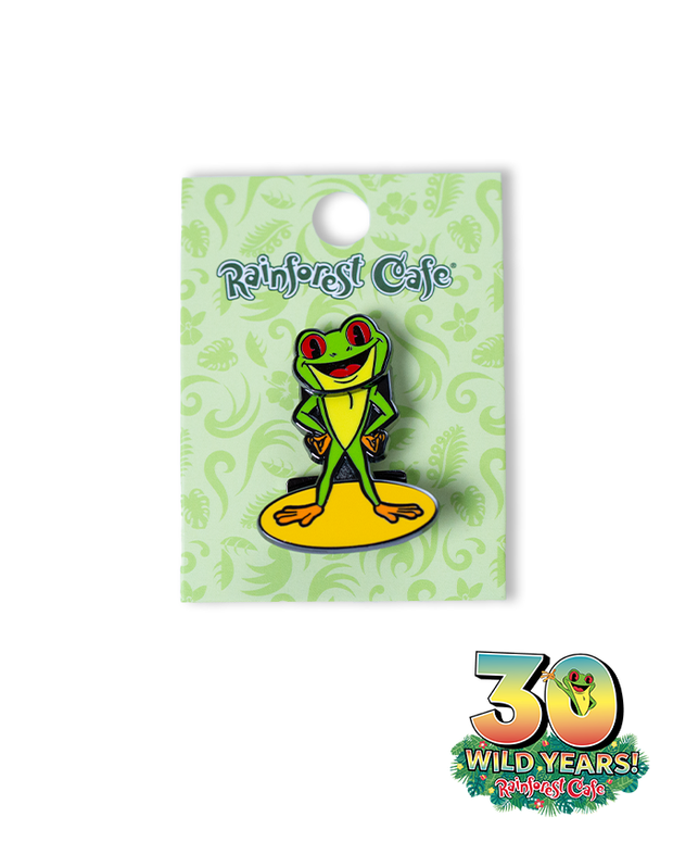 A colorful enamel pin featuring Cha Cha, the smiling frog mascot of Rainforest Cafe, standing with its arms crossed on a yellow platform. The pin is attached to a green card adorned with decorative patterns and the ‘Rainforest Cafe’ logo at the top. The card also celebrates ‘30 WILD YEARS!’ of Rainforest Cafe with a festive design at the bottom right corner.