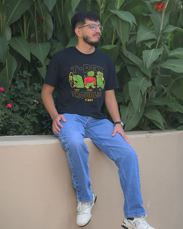A person wearing a black T-shirt with a colorful ‘T-REX TROUBLES’ graphic, blue jeans, and white sneakers, sitting on a ledge in front of green plants.