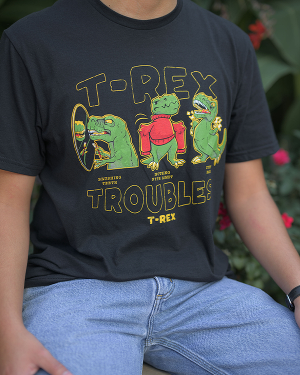 A close-up view of a model sitting in front of foliage, wearing a pair of blue jeans, white sneakers, and a black crewneck T-shirt with three green dinosaurs in funny situations, with the words "T-REX TROUBLES, T-REX" around it.