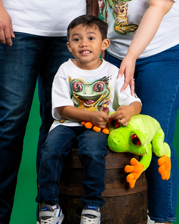 A child sits on an old-fashioned wooden barrel, wearing a white t-shirt with an animated frog design and blue jeans. Two adults stand beside the child, both in white t-shirts with animated designs and blue jeans. The child holds a green stuffed frog toy rests on the barrel next to him, all against a solid green background.