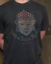 Close up of dark t-shirt featuring a colorful, flame-like design at the top and the inscriptions ‘Dwell on Positive Thoughts’ and ‘Yak & Yeti - Orlando’ stands with their face obscured by a grey square. The background suggests an indoor setting with wooden elements.
