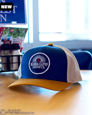 Blue and mustard trucker hat with white mesh on the back. The cap has a front round graphic with a small red anchor and  Bubba Gump Shrimp Co Restaurant and Market wording. The hat is on the table in one of Bubba Gump's restaurants. In the background, there is a window light. 