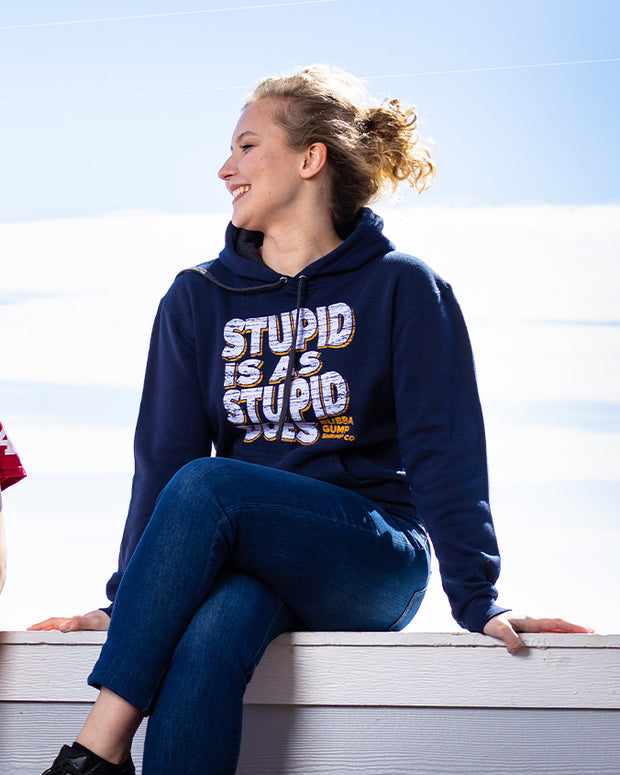 A girl is sitting on the ledge wearing a navy blue "Stupid is as Stupid Does" hoodie and smiling, looking to her right side. She also wears dark blue skinny jeans, and her hair is blond curly, and tied in a messy bun. There is a sunny blue sky in the background. 