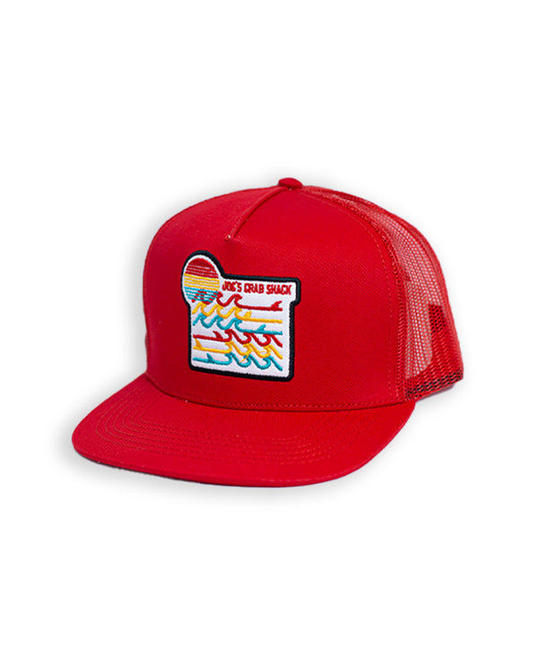A red baseball cap with an embroidered patch in the middle featuring "JOE'S CRAB SHACK" branding, and colorful waves and a sun. The background is plain white.