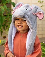 Small child smiling at the camera while wearing grey Tuki the Elephant plush hat and standing outside.