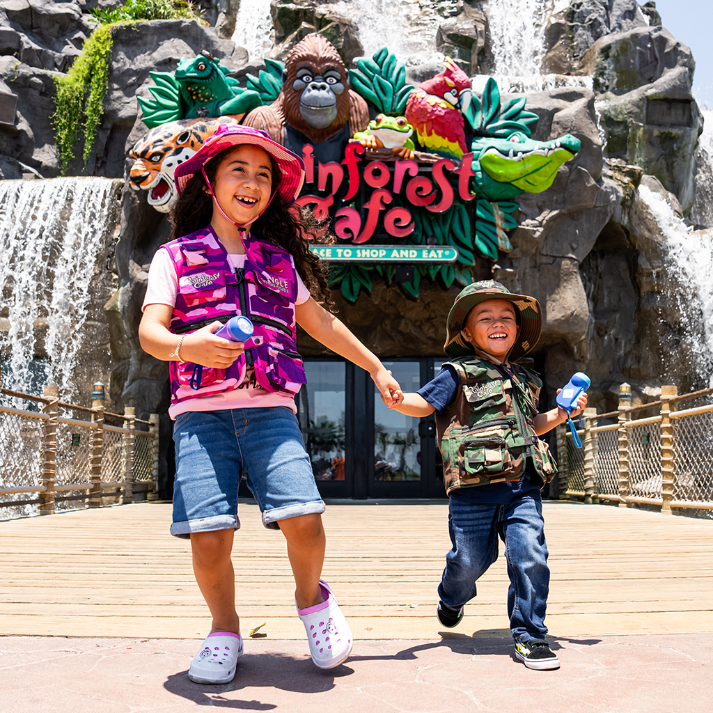 two kids in front of rainforest cafe restaurant sign, holding hands, wearing camo vest and hats.