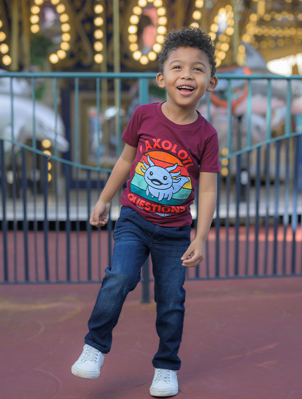 A child is pictured standing in front of a carousel, wearing a vibrant burgundy t-shirt with the playful graphic ‘I AXOLOTL QUESTIONS’. 