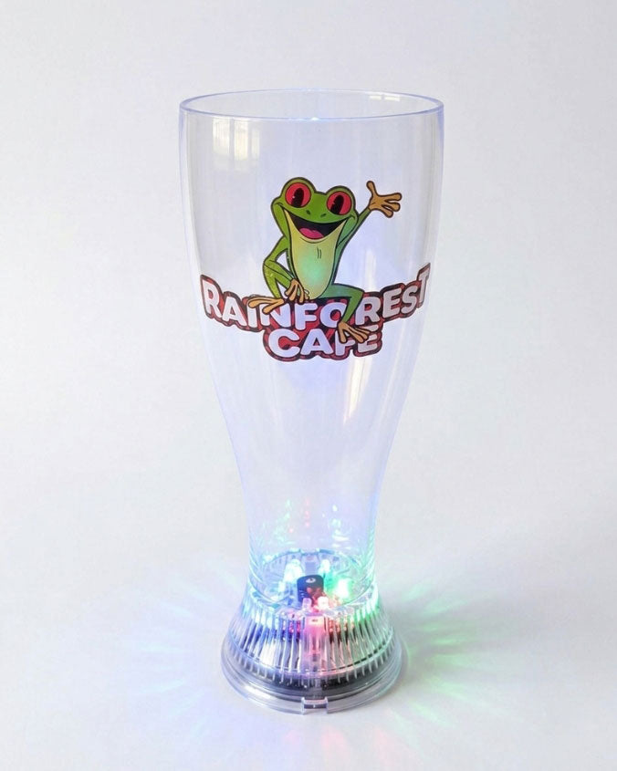 A clear plastic pint glass with a printed green frog and 'Rainforest Cafe' logo on the front, illuminated from within at the base with multicolor LED lights, set against a plain white background.