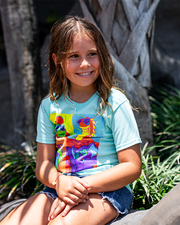 little girl sitting in front of plants and a palm tree wearing the 4 square kids tee and denim shorts.