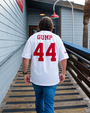 A man walks on a wooden path wearing a white jersey with "GUMP" and the number "44" on the back, heading towards a building.