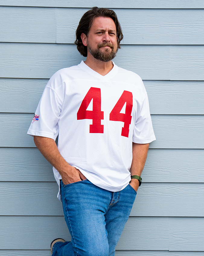 A person leans against a light blue wall, wearing a white jersey with red numbers "44" and blue jeans, exuding a casual vibe.