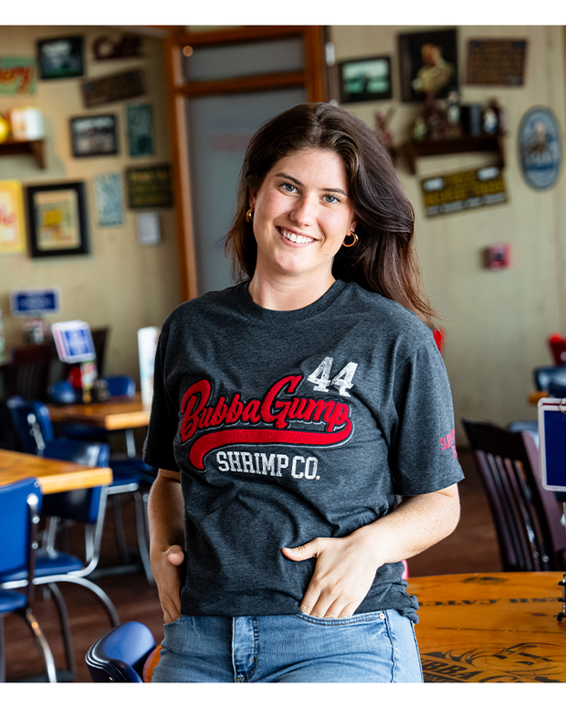 girl sitting on table in bubba gump restaurant wearing Charcoal tee with the word "Bubba Gump" in a red applique design. In white letters, under reads "Shrimp Co." and in white letters, the number 44 is on top left 