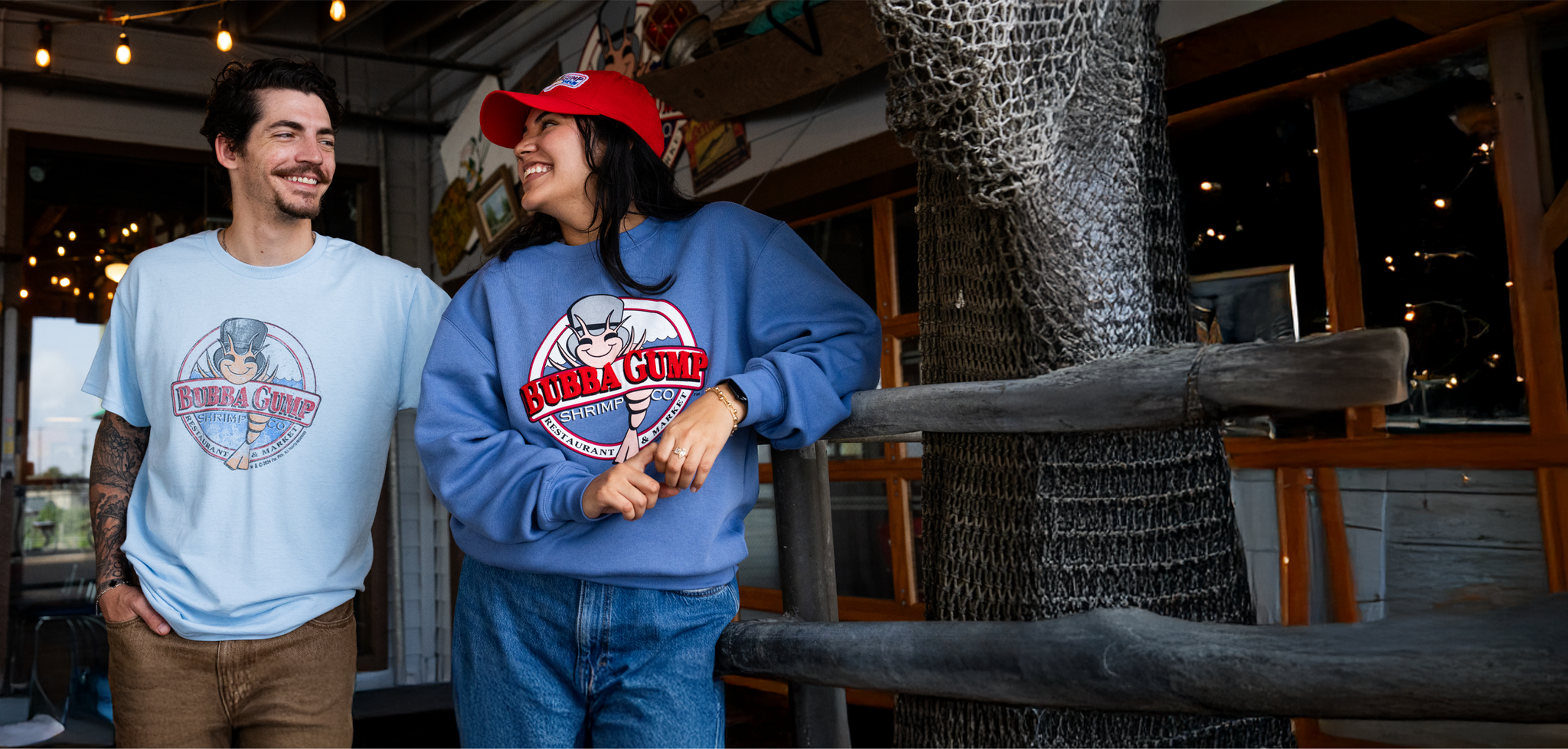 Two casually dressed individuals stand together, wearing branded Bubba Gump merchandise, with a rustic restaurant ambiance in the background.