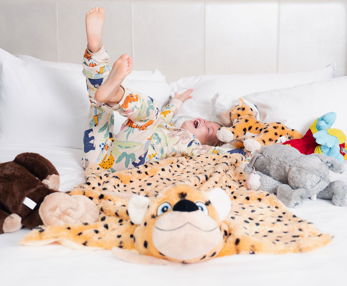A little girl wearing animal-printed pajamas playfully rolling on the white bed with a plush blanket and five other plush toys. 