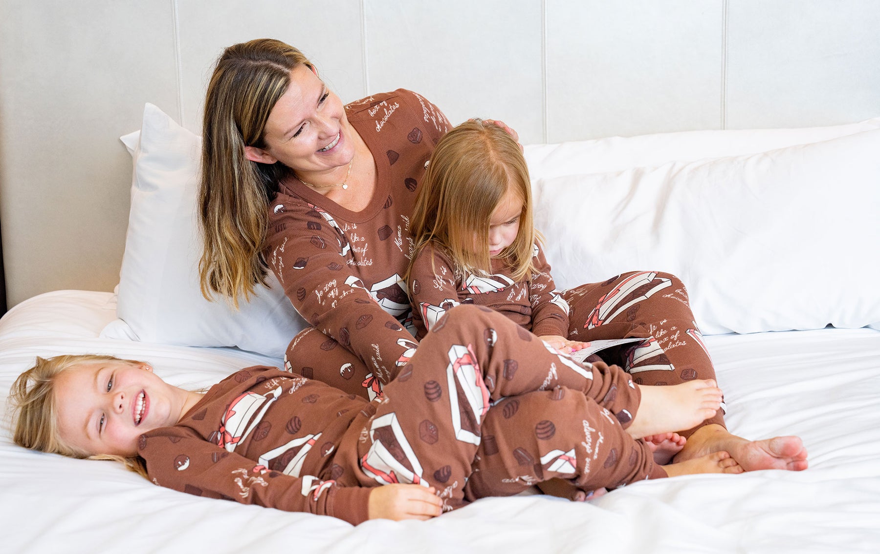 Mom and two daughters are laughing and lying on the white bed wearing matching brown pajamas. 