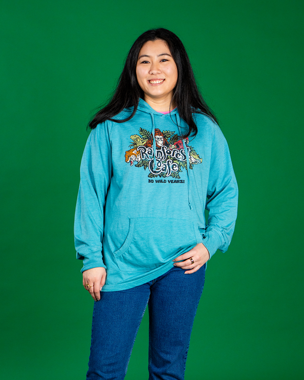 A person wearing a teal hoodie with the ‘Rainforest Cafe 30 WILD YEARS!’ logo, standing against a green background. 