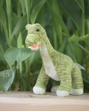 A plush green brachiosaurus toy with a playful expression, standing on a ledge in front of vibrant green plants.