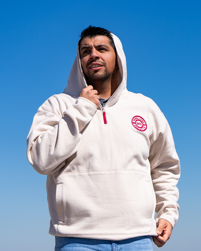 male model wearing cream hooded jacket. He is looking to the side, with his right hand on collar. jacket has kangaroo pockets, quarter zip with red tag on zipper, and red emblem on left chest.